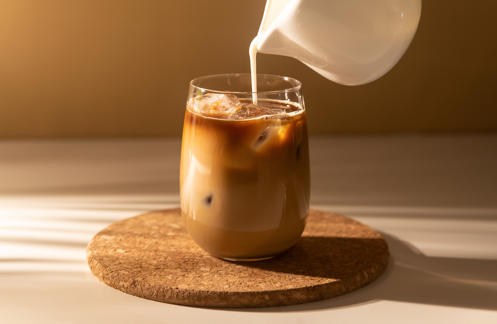 Milk cream is poured into a iced cold brew coffee. Coffee cold cocktail drink with ice and milk in morning sun light.