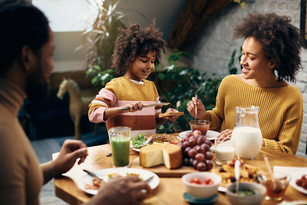 Happy black girl spreading butter on toasted bred while eating breakfast with her parents in dining room.