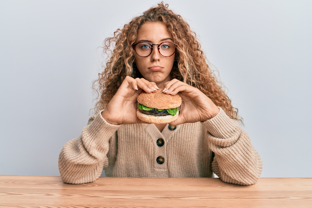 Beautiful caucasian teenager girl eating a tasty classic burger depressed and worry for distress, crying angry and afraid. sad expression.
