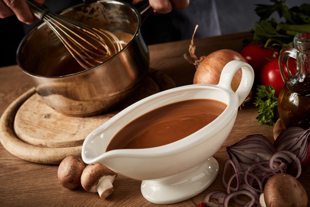 Sauce boat filled with rich brown spicy gravy viewed high angle on a kitchen table with assorted fresh vegetable ingredients