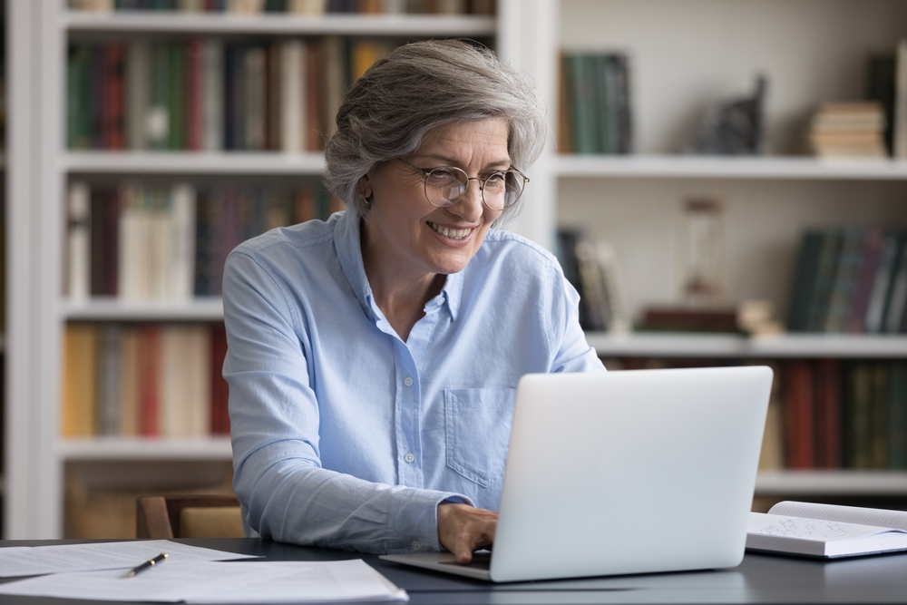 Happy successful smart old senior 60s woman in eyeglasses looking at laptop screen, working online from home, enjoying distant study, communicating in internet or web surfing information remotely.