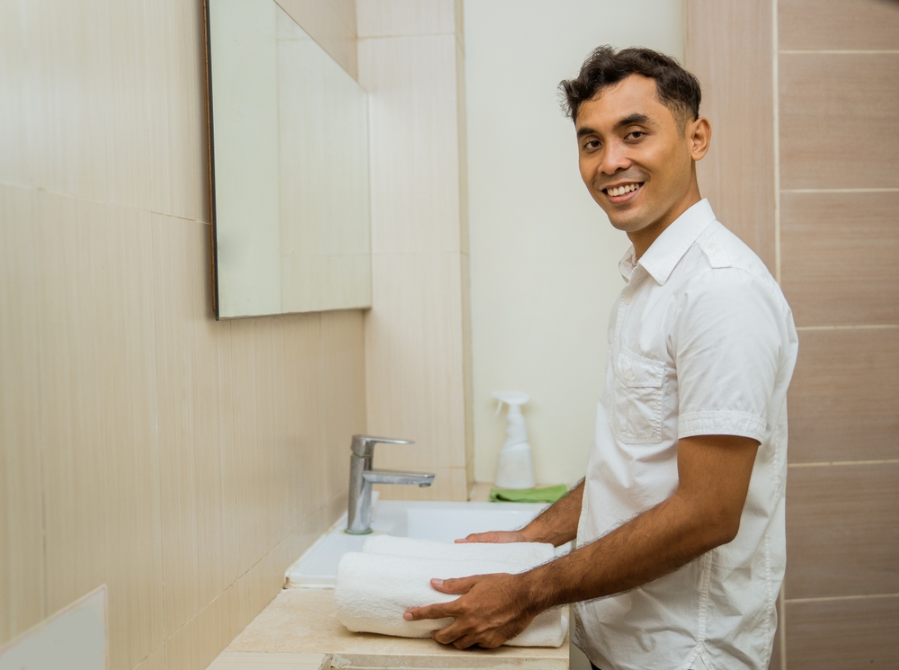 smiling asian male janitor putting towel beside sink in bathroom
