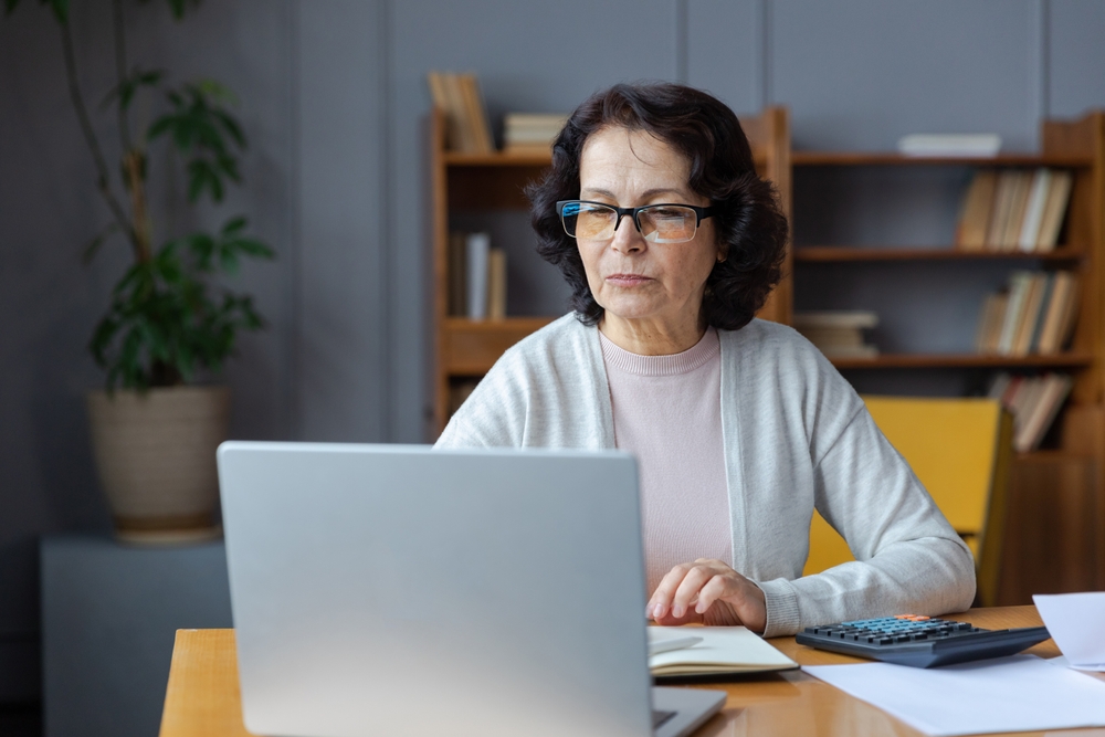 Middle aged senior woman sit with laptop and paper document. Pensive older mature lady reading paper bill pay online at home managing bank finances calculating taxes planning loan debt pension payment