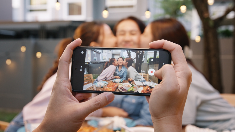 Young adult asia people hug cuddle kiss love care for mom taking photo selfie video on mobile phone camera at home picnic dining fun night party dine table. Relax older mum smile enjoy warm time meal.
