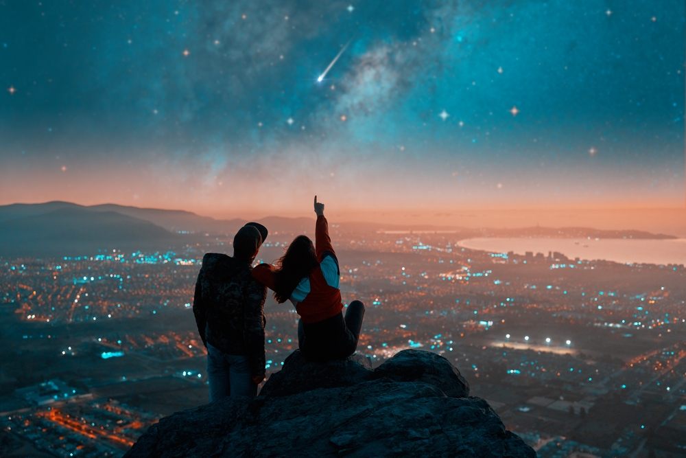 silhouettes of a couple sitting on the top of the mountain looking and pointing out at shooting star and milky way over the city lights on the horizon