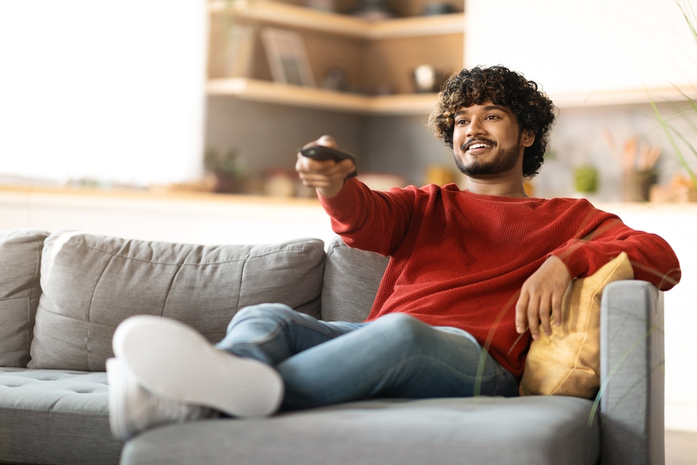 Home Relax. Handsome Indian Guy Sitting On Couch And Switching Tv Channels, Smiling Young Eastern Man Holding Remote Controller, Relaxing On Comfortable Sofa In Living Room, Copy Space
