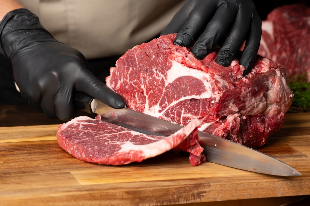The butcher wearing black gloves cuts the meat with knife on wooden cutting board.Close-up piece of raw meat with a chef's knife on a dark background.