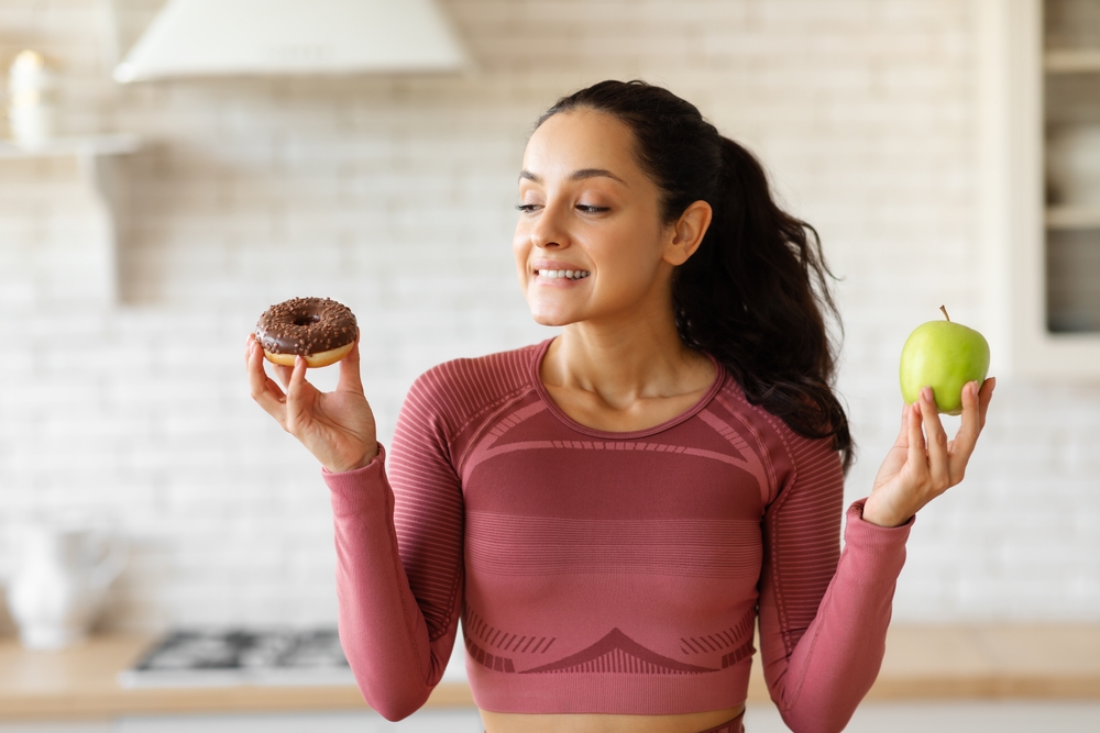 Athletic woman in activewear choosing apple fruit or donut struggling from sugar cravings during diet, standing in kitchen setting. Unhealthy vs healthy food choice concept