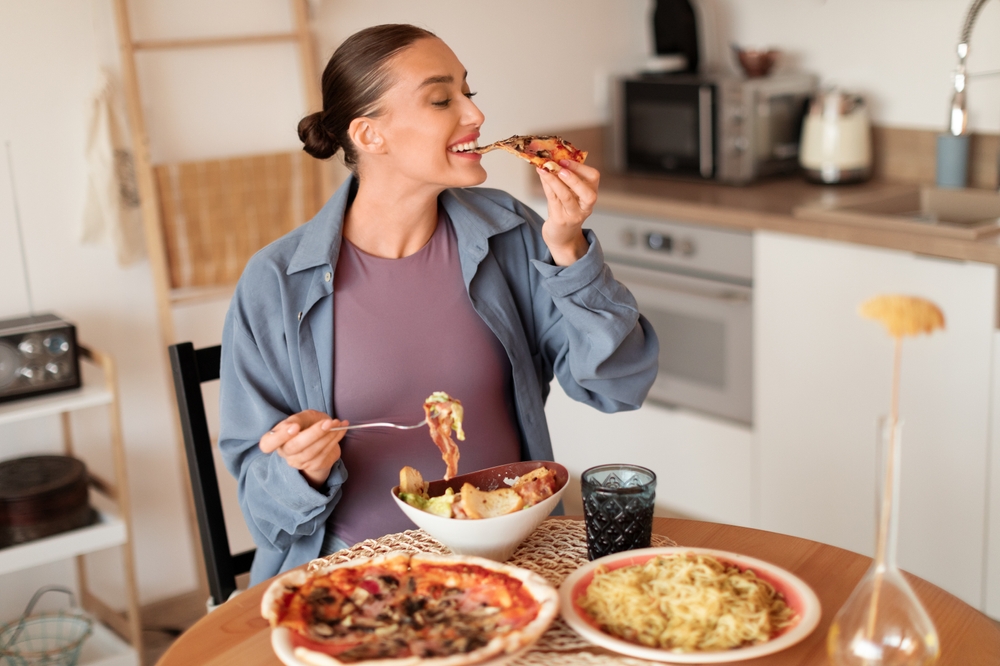 Expecting mother in relaxed attire relishes pizza and pasta dishes, signifying diverse cravings during pregnancy in a warm domestic environment