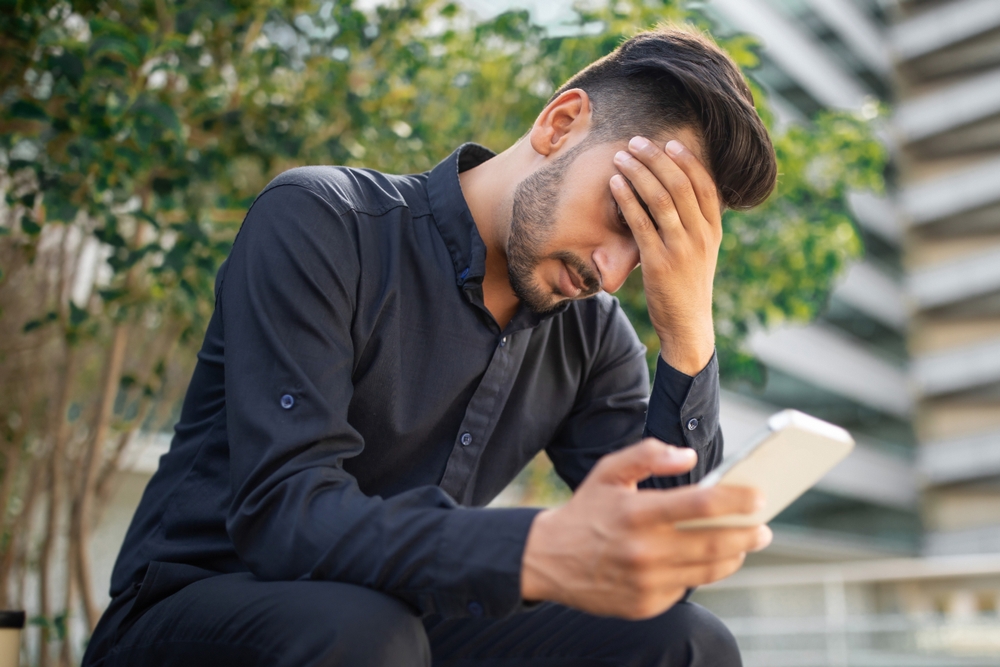 Distressed young middle eastern businessman covers face with hand, receiving bad news on phone sitting in urban area outdoor, stressed entrepreneur having headache after doomscrolling
