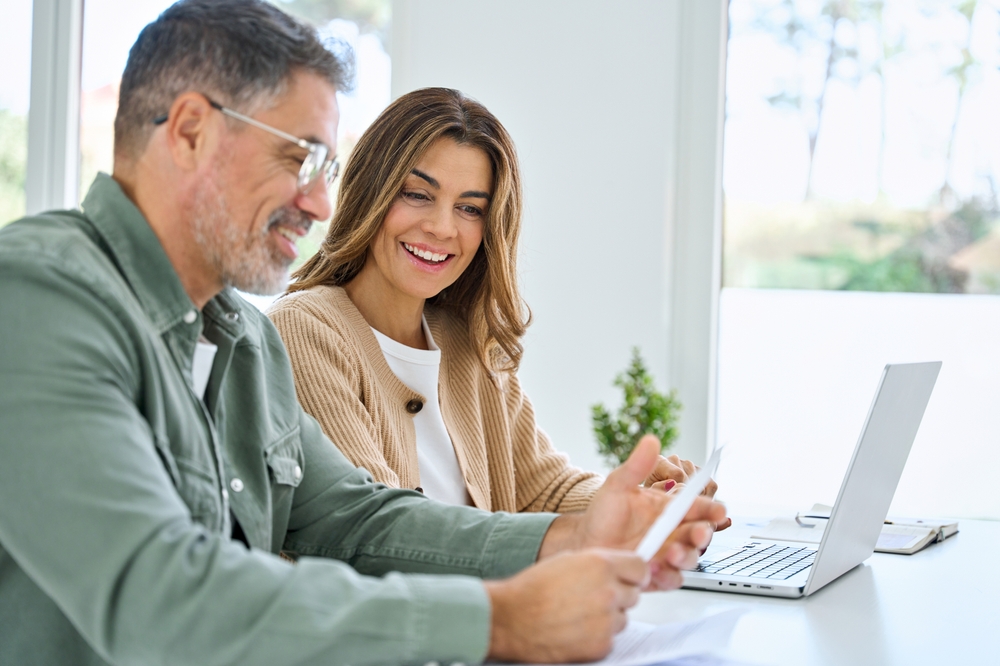 Happy middle aged mature couple using laptop counting taxes refund receipts to save money at home. Old man and woman paying bills online planning financial budget savings calculating payment at table.
