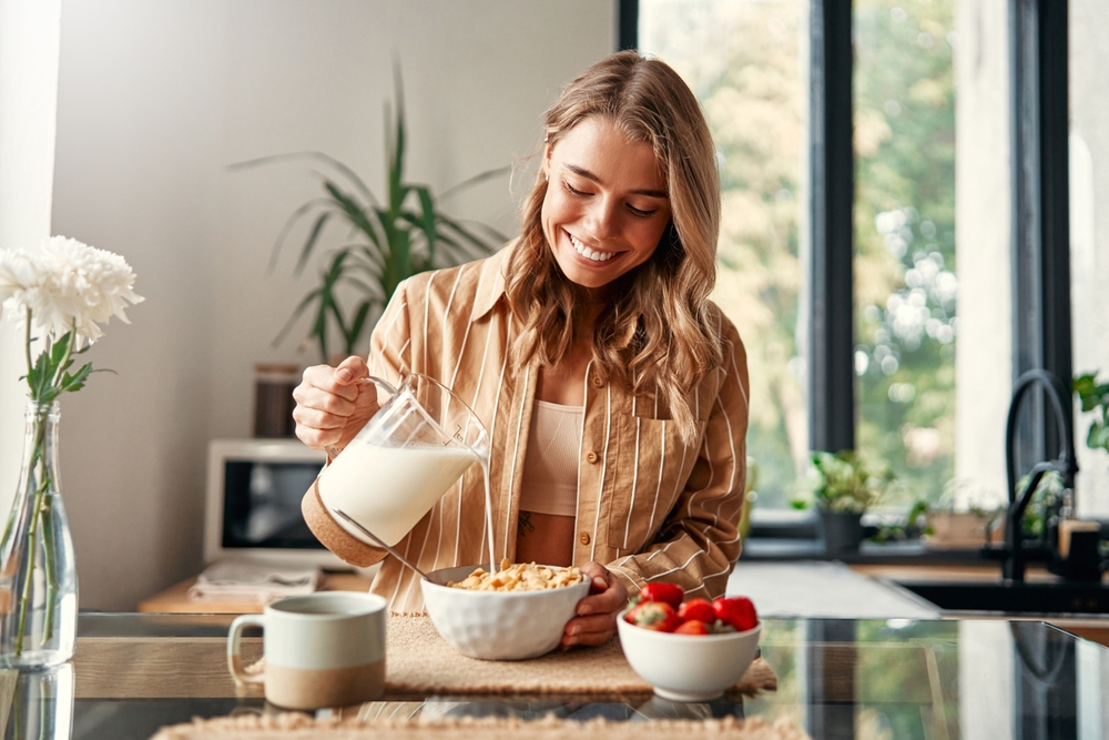 A woman preparing herself breakfast, pouring milk into a plate with cereal, strawberries and a cup of coffee standing nearby. A woman is having breakfast in a cozy kitchen.