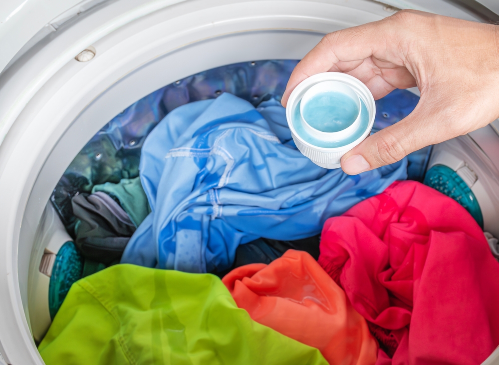 Hand holding laundry detergent with colorful clothes in washing machine

