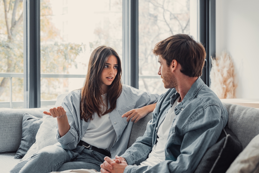 Young happy couple having fun talking laughing relaxing at home on couch, boyfriend embracing girlfriend telling funny joke sitting on sofa, humor in relationships, enjoying weekend together
