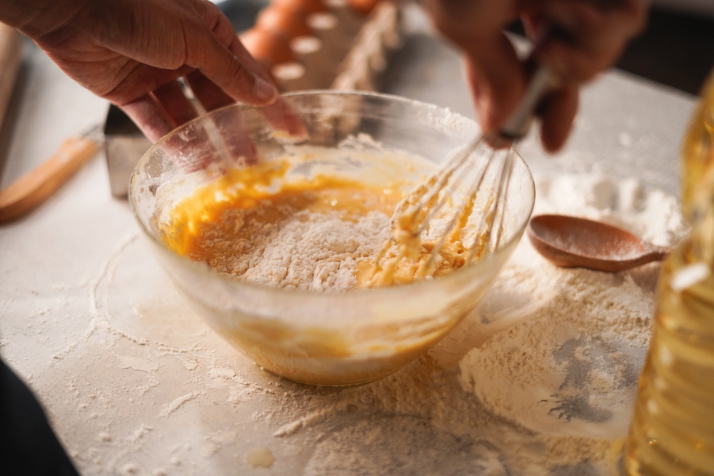 Close-up of hands whisking flour and eggs in bowl during baking preparation. Ingredients on table. authentic baking scene. homemade baking, cooking process and culinary activities
