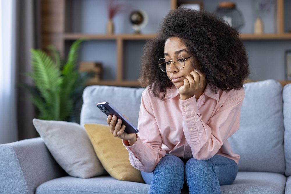 Young woman sitting on a sofa looking bored and uninterested at her phone. She's feeling lonely and disconnected while spending time at home. Concept of boredom, loneliness, and disinterest.
