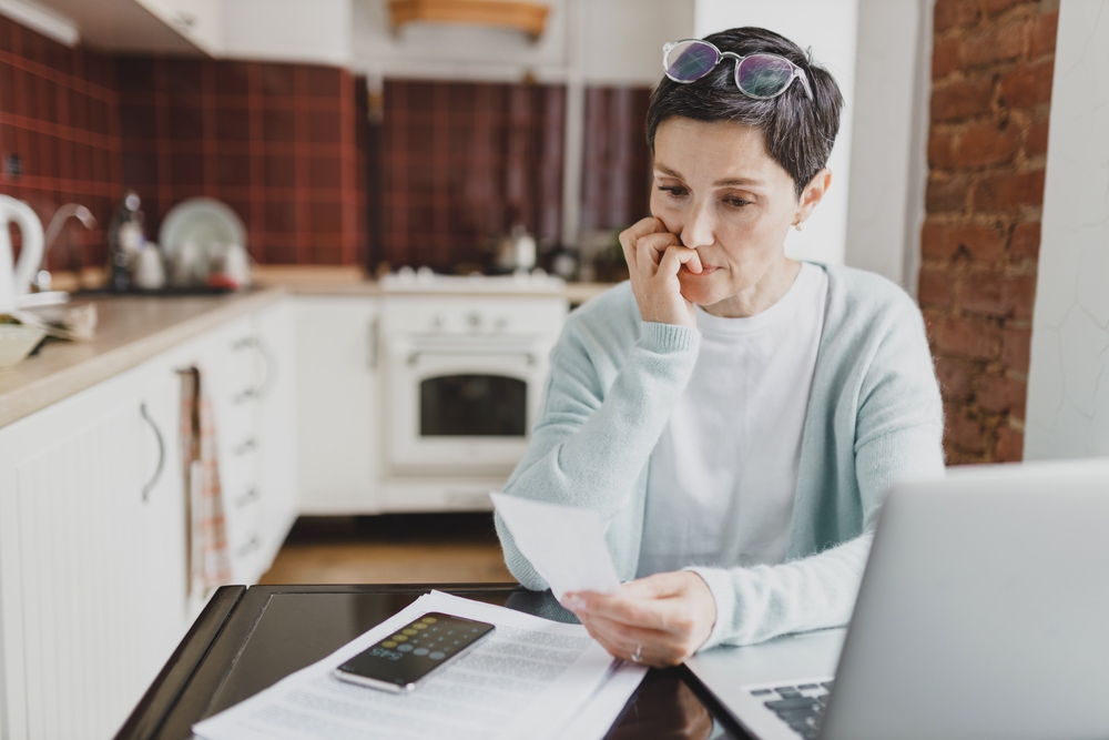 Selective focus on worried anxious female of 60s biting nails holding utility bill in hands sitting at kitchen table, trying to calculate expenditures, using laptop to pay and smartphone calculator