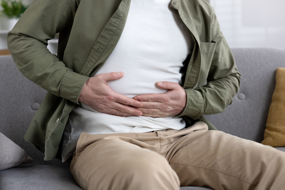 Man sitting on couch experiencing abdominal pain holding stomach. Wearing casual clothing and showing visible discomfort. Concept of stomachache, digestive issues, or health concerns.
