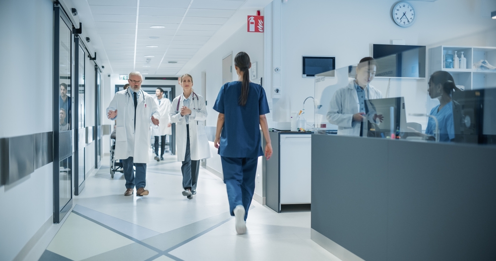 Diverse Male and Female Medical Professionals Navigate a Busy Hospital Corridor and Registration Area, Having Conversations About Healthcare. Male Nurse Assists a Patient in a Wheelchair
