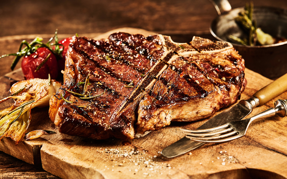 Succulent grilled large t-bone steak garnished with herbs, tomato and salt with fork and knife beside it on cutting board
