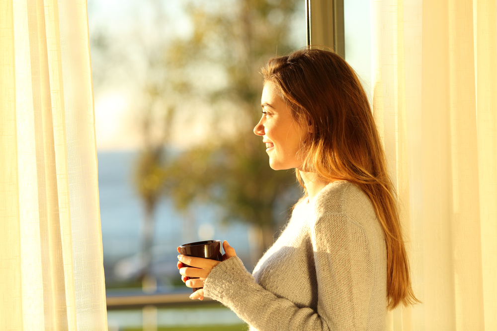 Side view portrait of a single relaxed girl looking the sea outside through a window and holding a coffee mug at sunset in the living room at home
