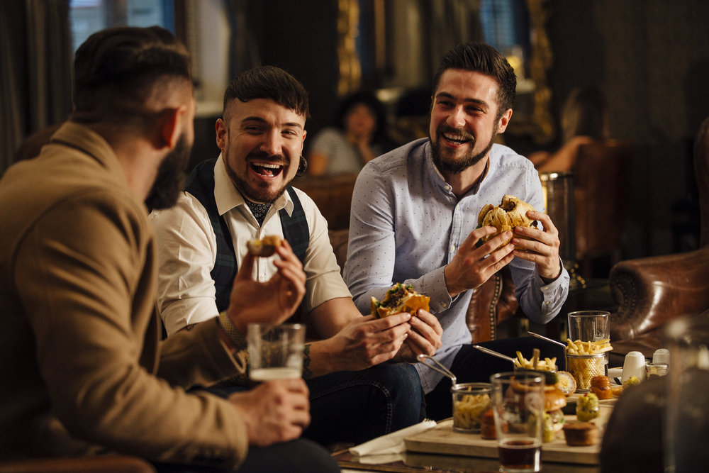 Three men are sitting together in a bar/restaurant lounge. They are laughing and talking while enjoying burgers and beer.
