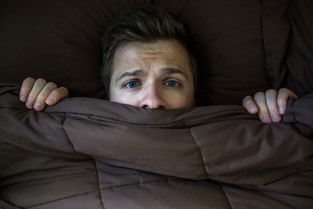 Caucasian young man hiding in bed under the blanket at home. He is stressed and tries run away from problems