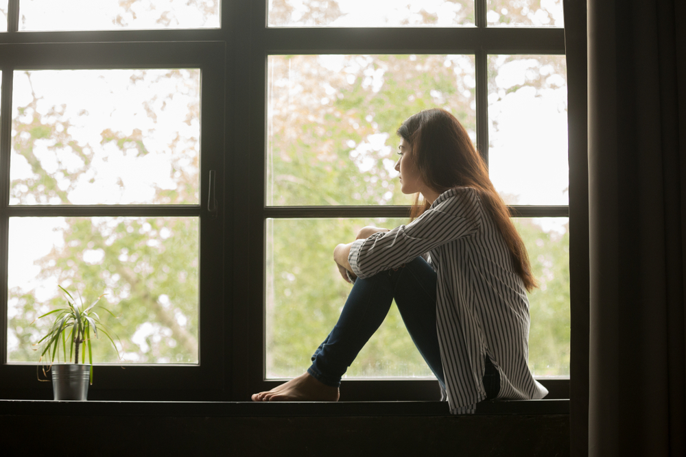 Thoughtful girl sitting on sill embracing knees looking at window, sad depressed teenager spending time alone at home, young upset pensive woman feeling lonely or frustrated thinking about problems
