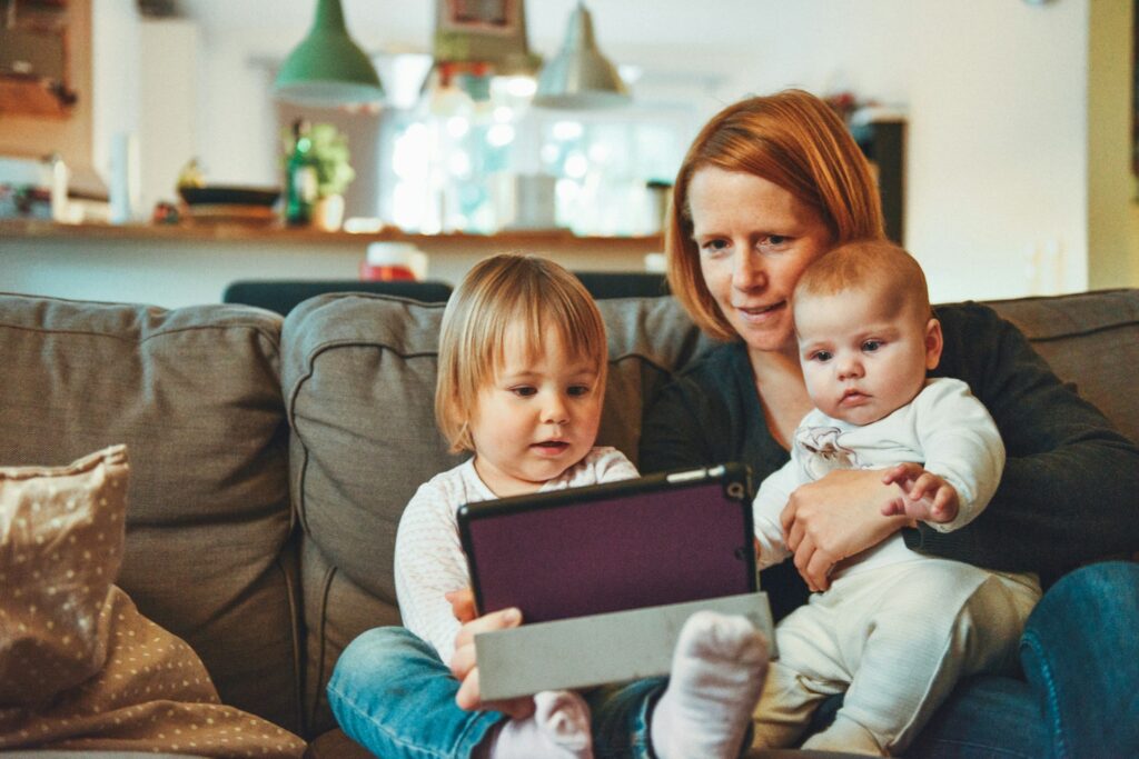 two babies and woman sitting on sofa while holding baby and watching on tablet
