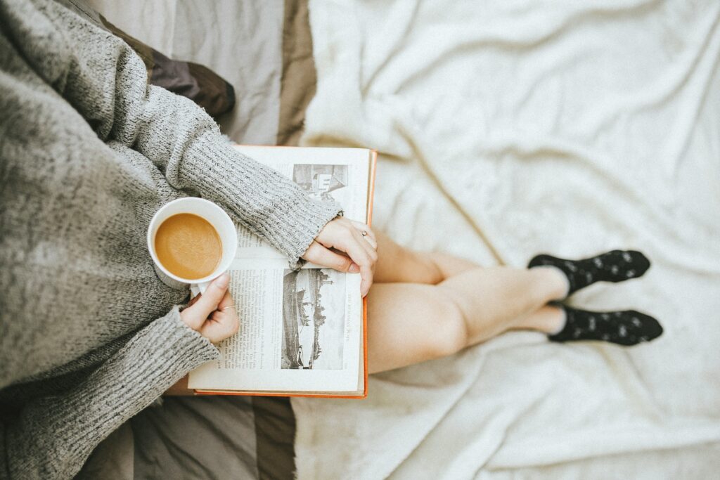 woman holding a cup of coffee at right hand and reading book on her lap while holding it open with her left hand in a well-lit room
