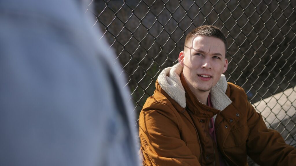 man wearing brown jacket sitting near gray link fence
