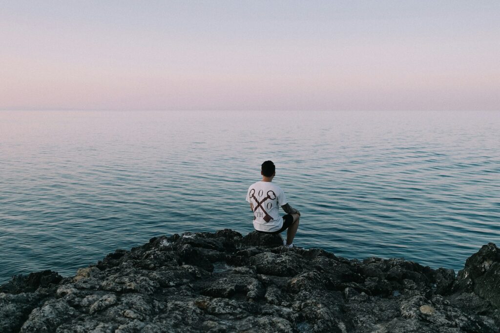 man in white t-shirt sitting on rock by the sea during daytime
