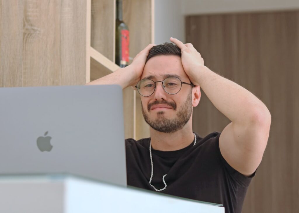 Man with hands pressed to head looking overwhelmed while working at laptop.