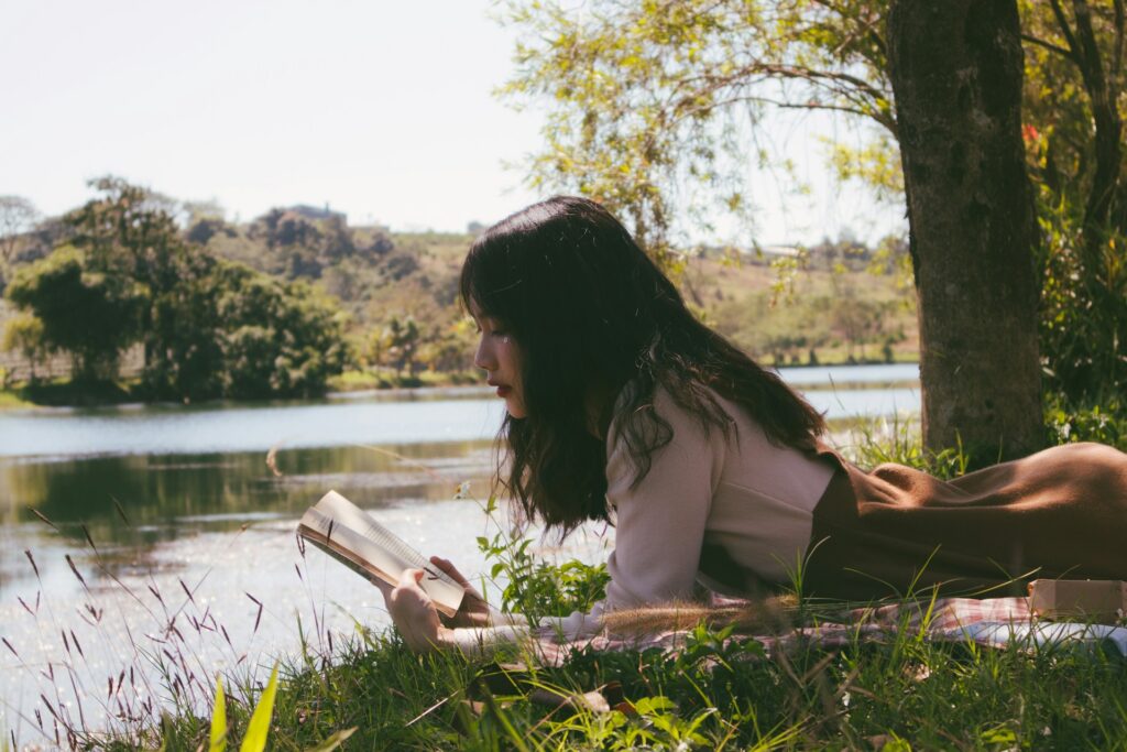 woman reading book and lying forward on sheet on grass beside body of water during day
