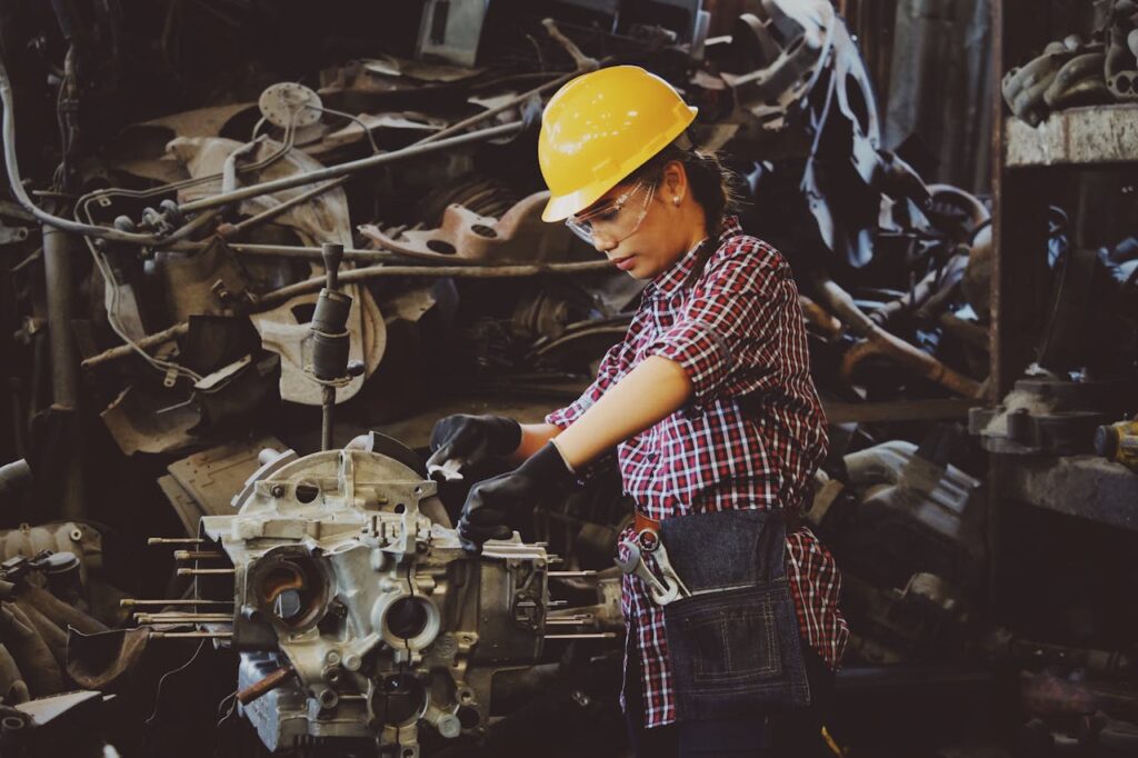 Woman Wears Yellow Hard Hat Holding Vehicle Part
