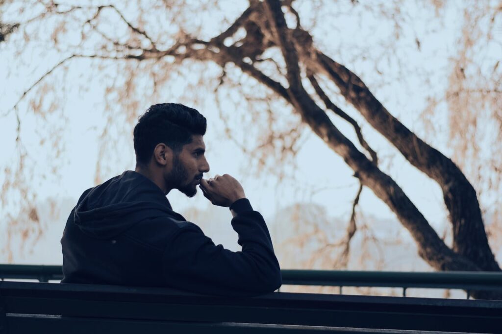 Man in Black Hoodie Sitting on Bench Near Green Trees
