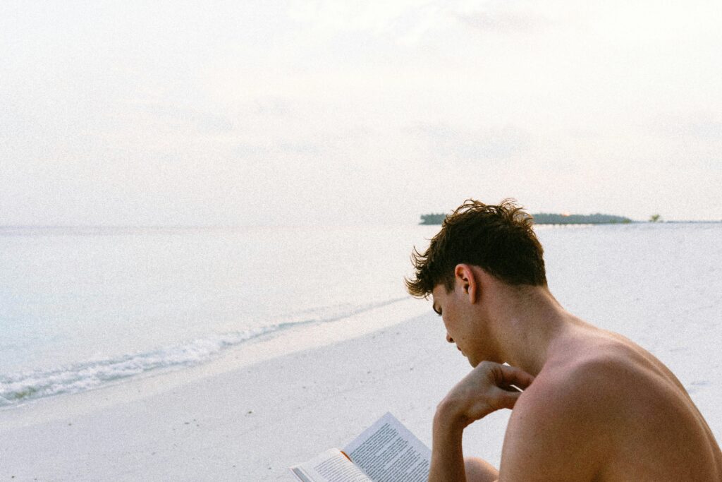 Topless Man Reading Book While Seating at Beach
