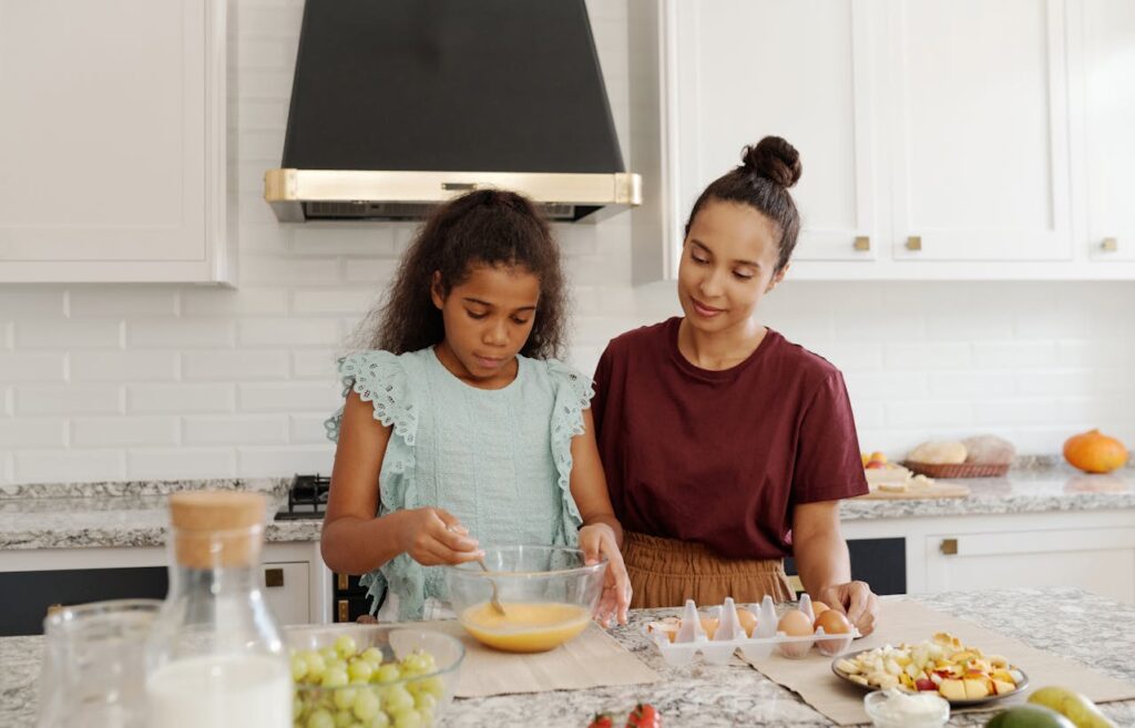 A Girl Baiting the Egg on a Bowl while Her Mother is Standing Beside Her
