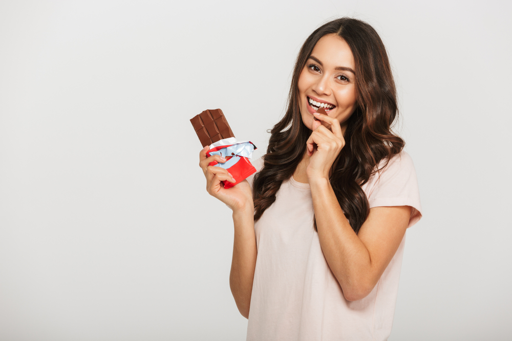 Portrait of a happy young asian woman eating chocolate bar isolated over white background
