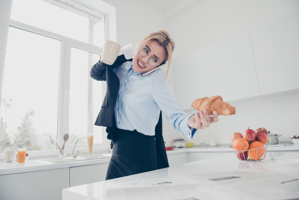 Adorable busy attractive charming beautiful smiling lady office executive worker wearing spectacles in hurry early in the morning talking on the phone having a drink and croissant in kitchen
