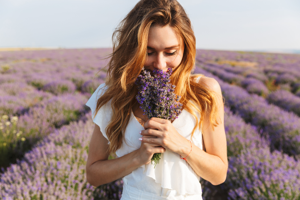 Photo of caucasian young woman in dress holding bouquet of flowers while walking outdoor through lavender field in summer
