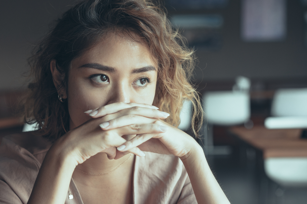 Closeup of sad young Asian woman at cafe covering nose and mouth with clasped hands and staring into vacancy. Upset manager thinking over mistakes. Failure and regret concept