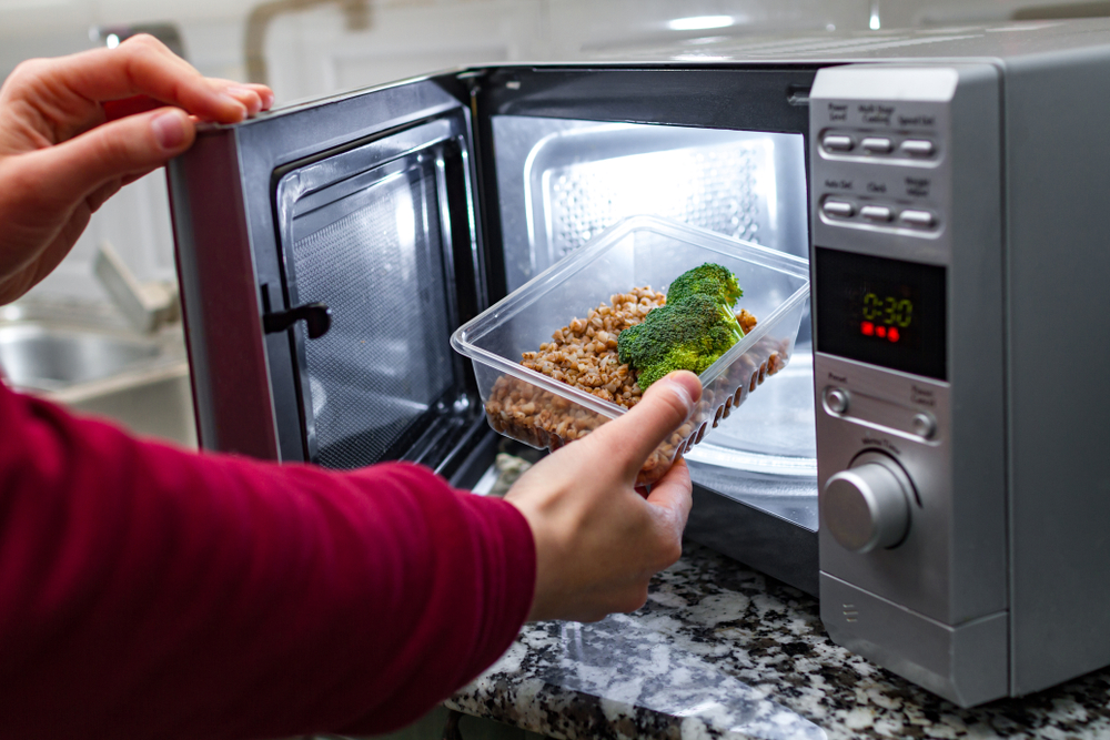 Using the microwave oven to heat food. Woman's hand puts plastic container with broccoli and buckwheat in the microwave