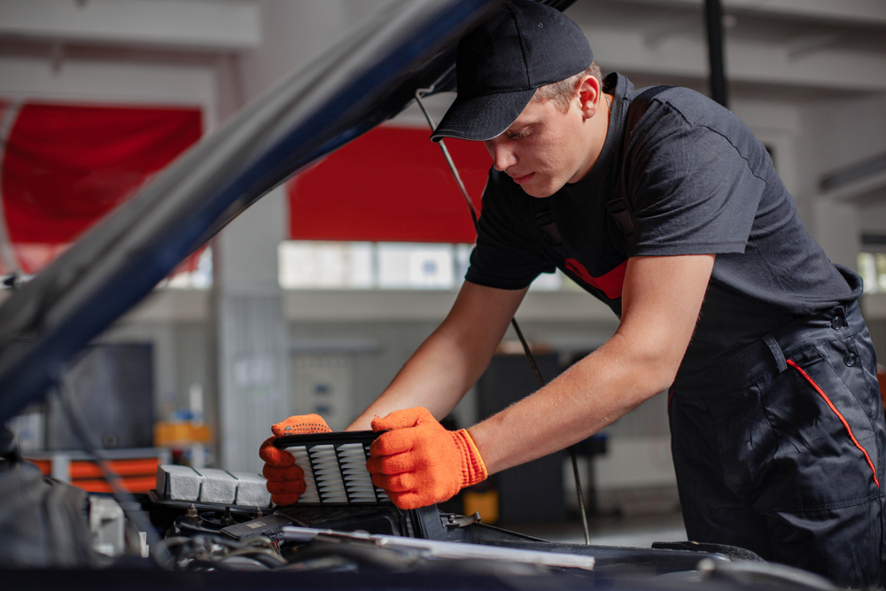 Mechanic is changing an air filter. Car hood is open.
