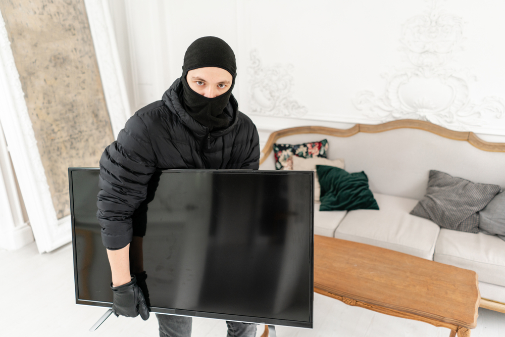 Thief looking at the camera, with black balaclava stealing modern expensive television. Luxury apartment with stucco. Man burglar stealing tv set from house