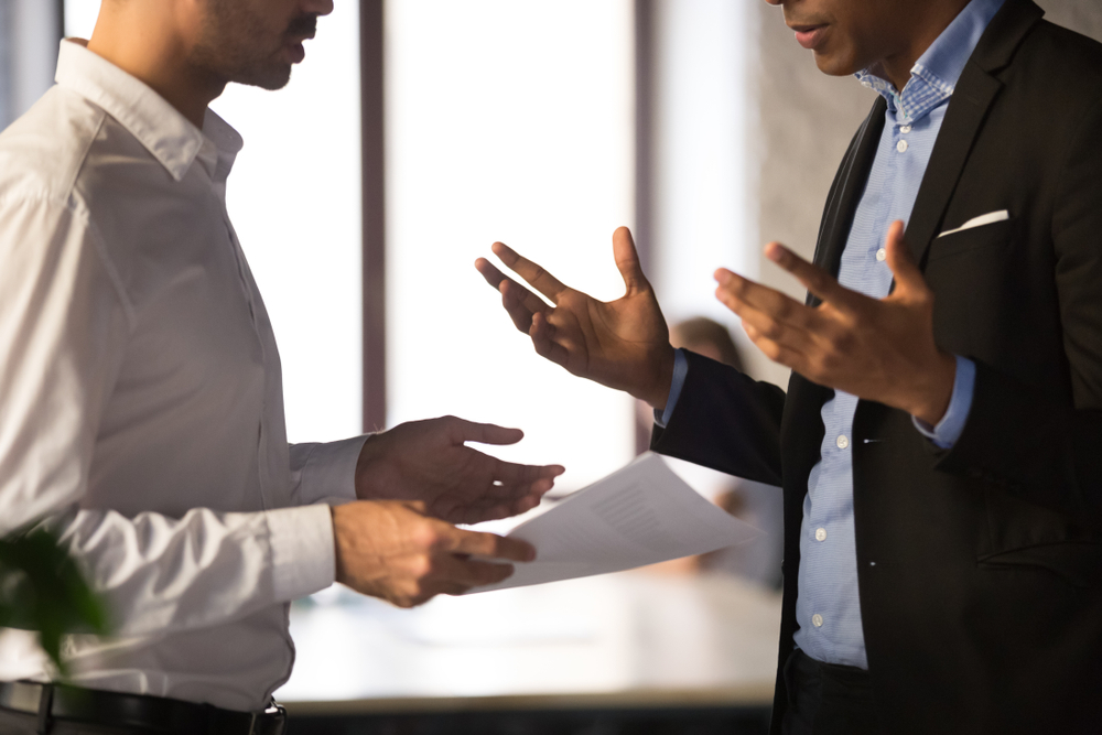 Close up executive manager dissatisfied by African American employee work results, holding financial report with stats, colleagues arguing, discussing business failure, partners disputing at work
