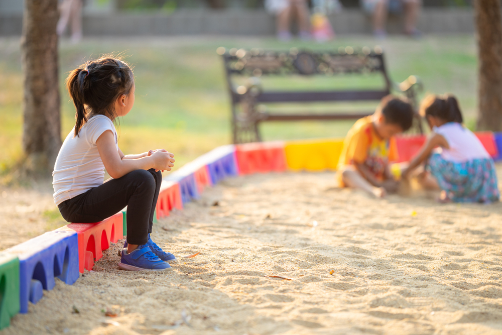 Little girl sitting lonely watching friends play at the playground.The feeling was overlooked by other people.Concept child shy.
