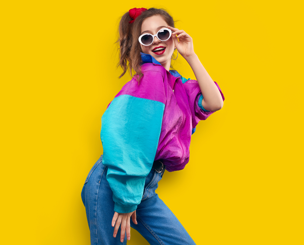 Cool teenager. Fashionable DJ girl in colorful trendy jacket and vintage retro sunglasses enjoys style of 80s - 90s vibes. Teenager Girl at disco party. Young fashion model on yellow color background.
