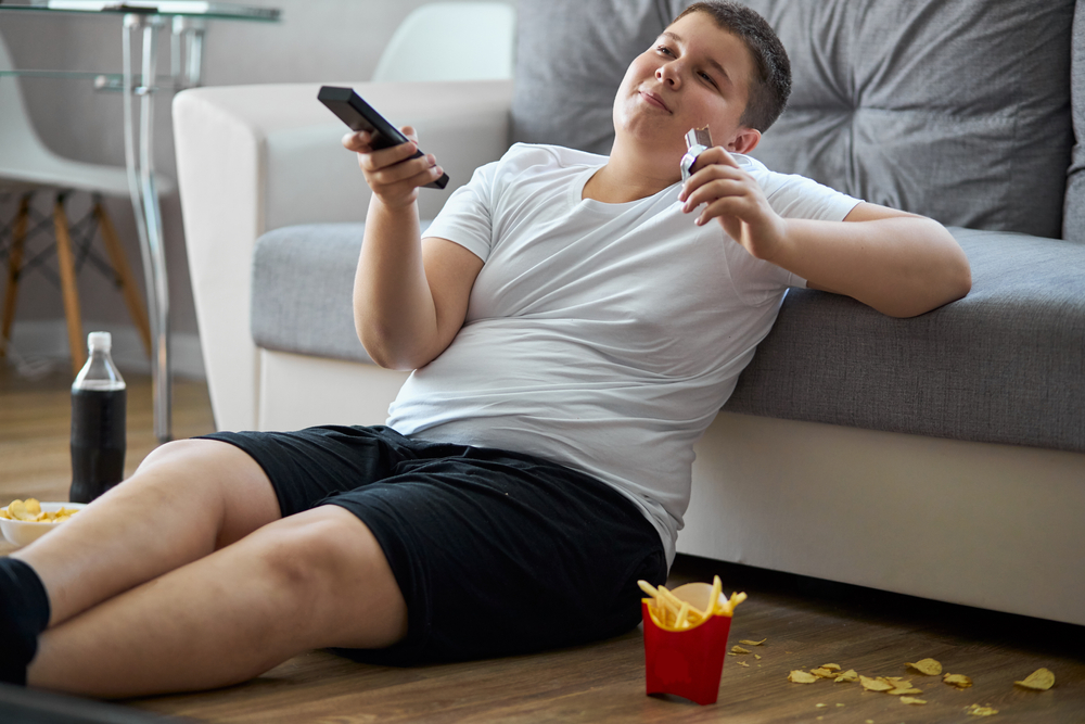 overweight boy eat junk food while watching tv alone at home, sit on the floor with french fries, carbonated drink, chips. enjoy unhealthy lifestyle