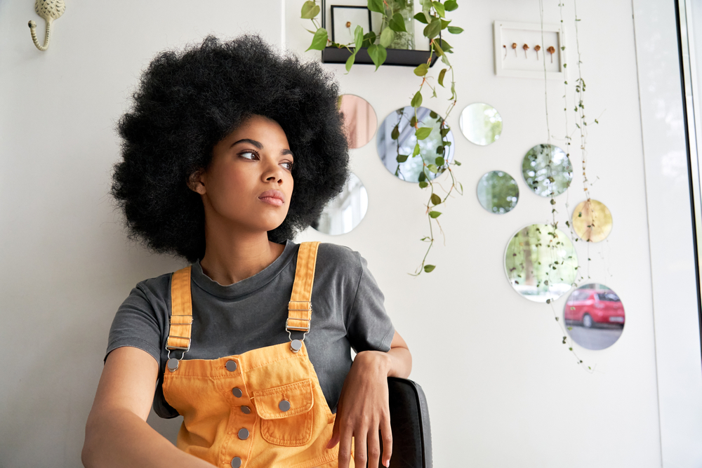 Young thoughtful pensive stylish African American gen z female hipster woman with afro hair looking through window thinking reflecting sitting lost in thoughts at home, in modern cafe alone indoors.
